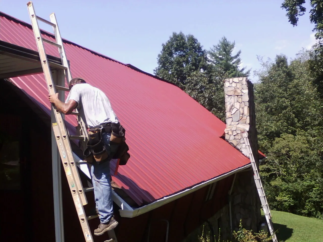 Roofer on a ladder beside a red metal roof and stone chimney — Soffit & Fascia Repair by Roofing Brothers in Armbrust